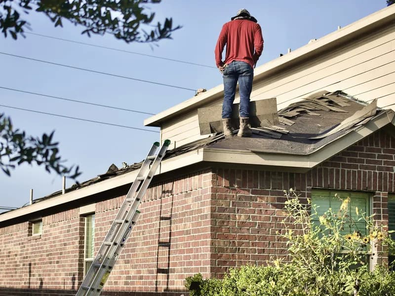 Roofer standing on a partly torn-off shingle roof on a brick home, ladder against the wall