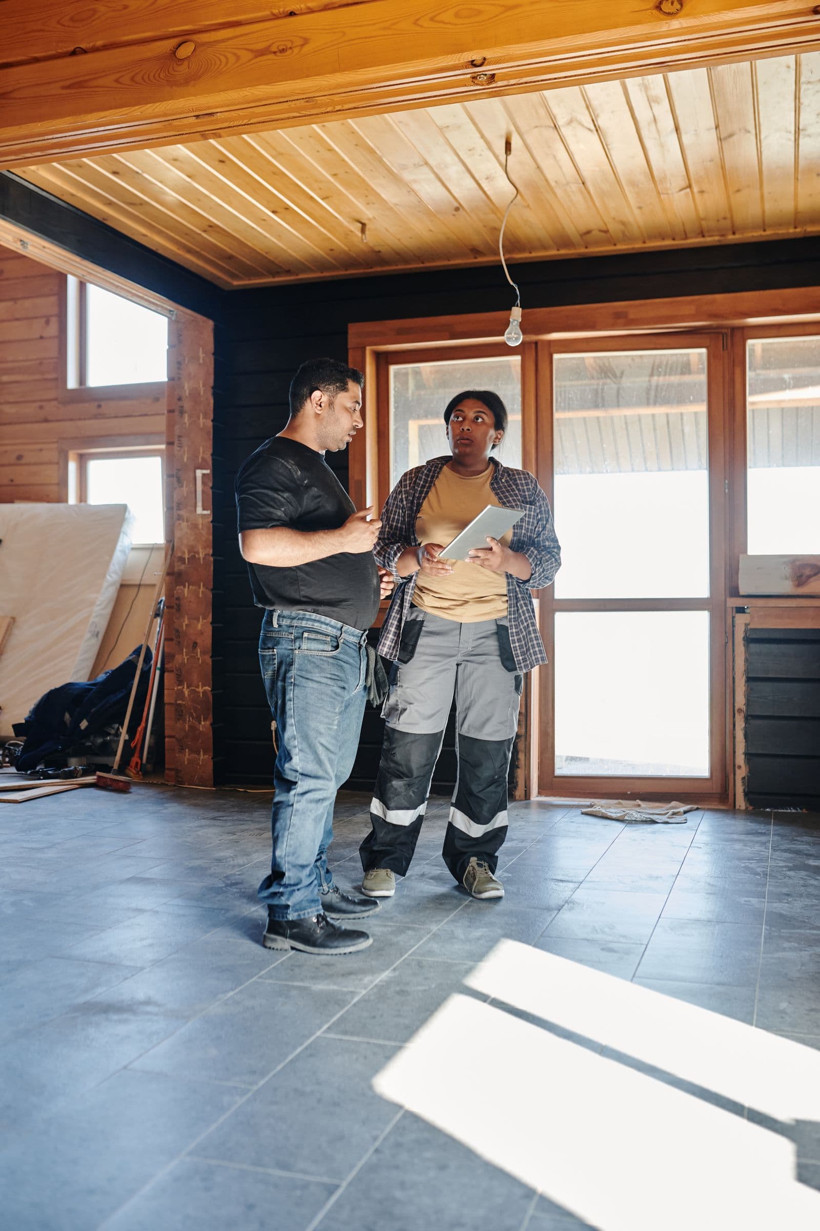 Two people in work clothes reviewing plans on a tablet inside a house under renovation
