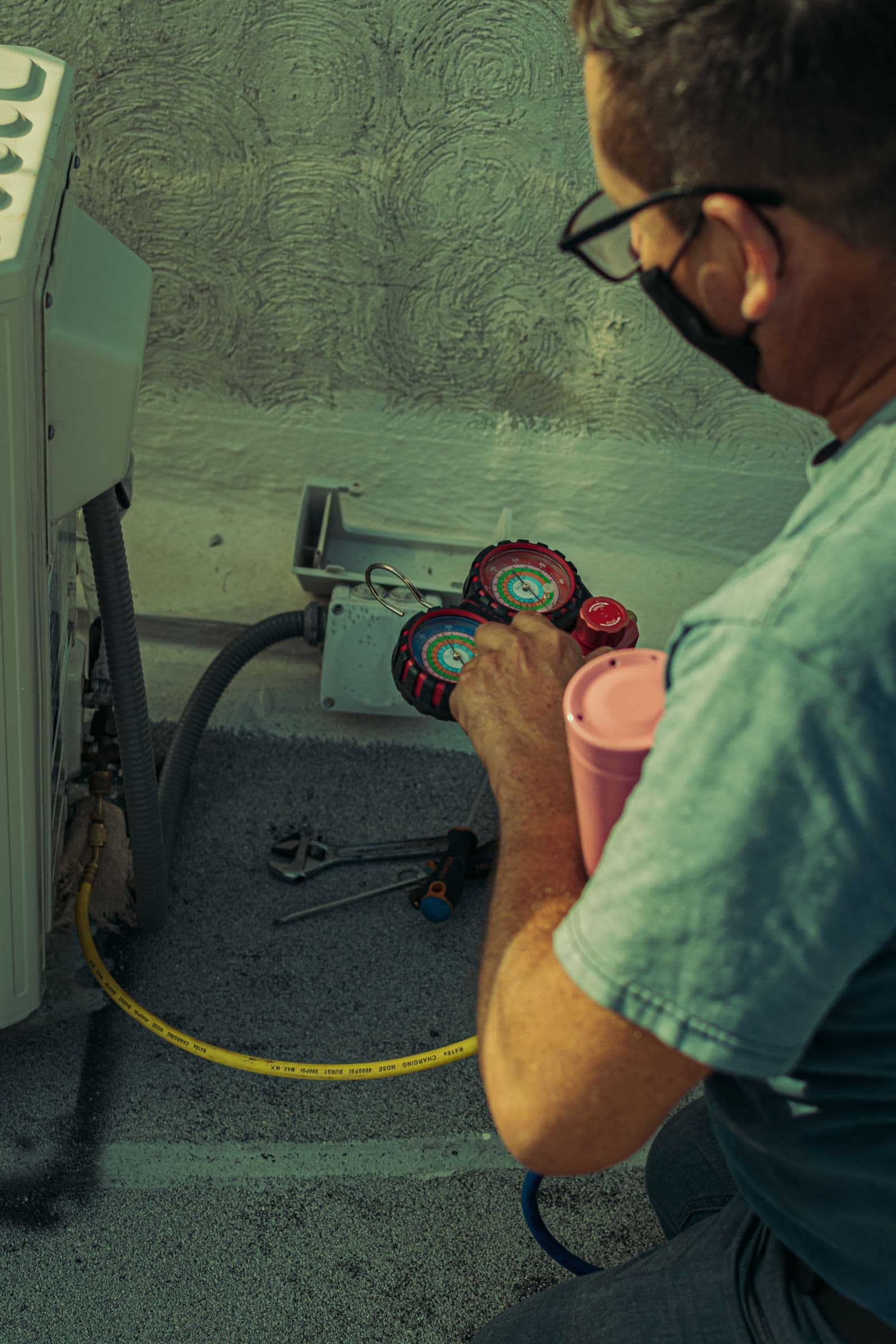 An HVAC technician kneeling at an outdoor condensing unit holding refrigerant manifold gauges