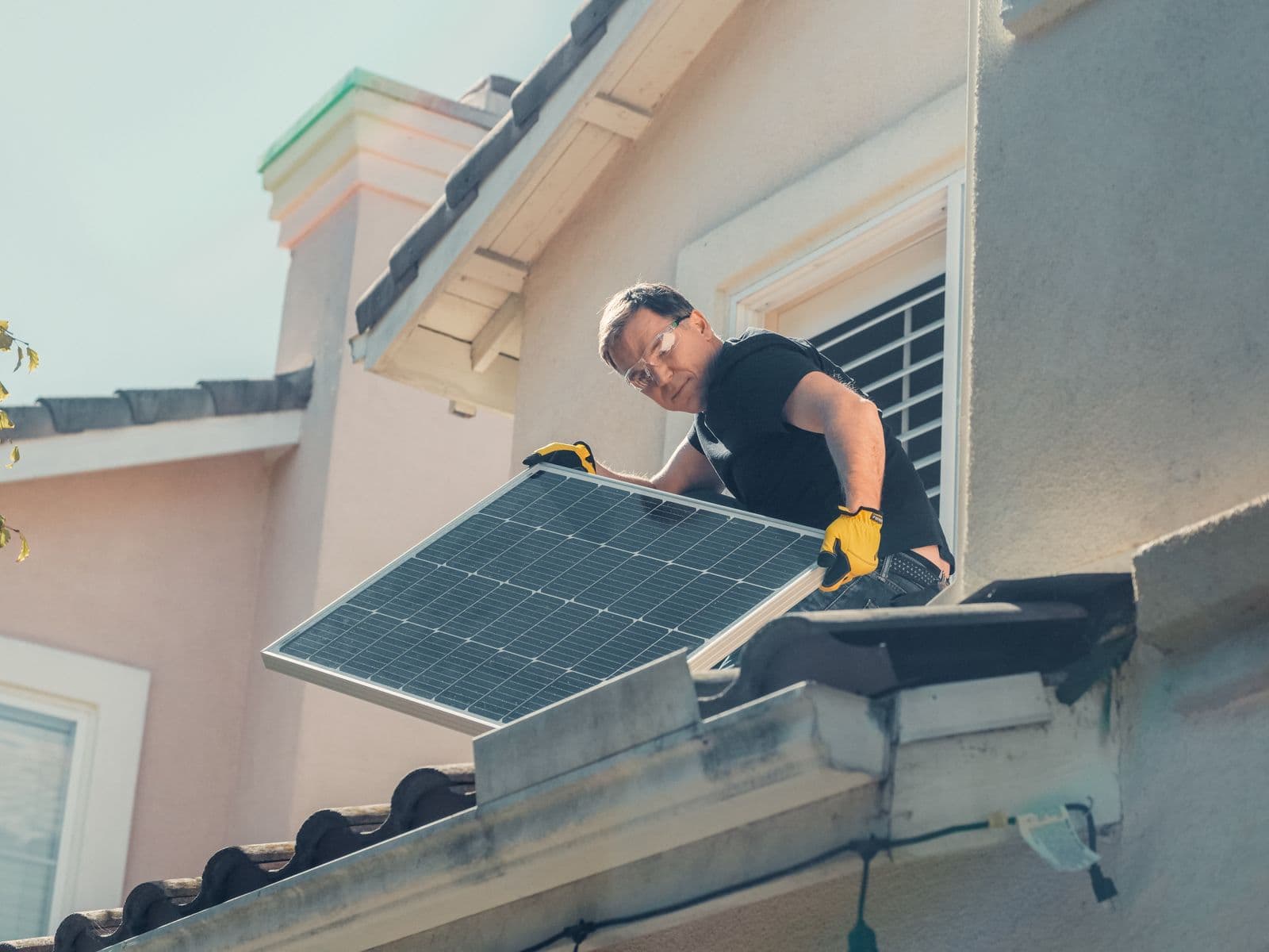 An installer in safety glasses positioning a solar panel on a tiled residential roof