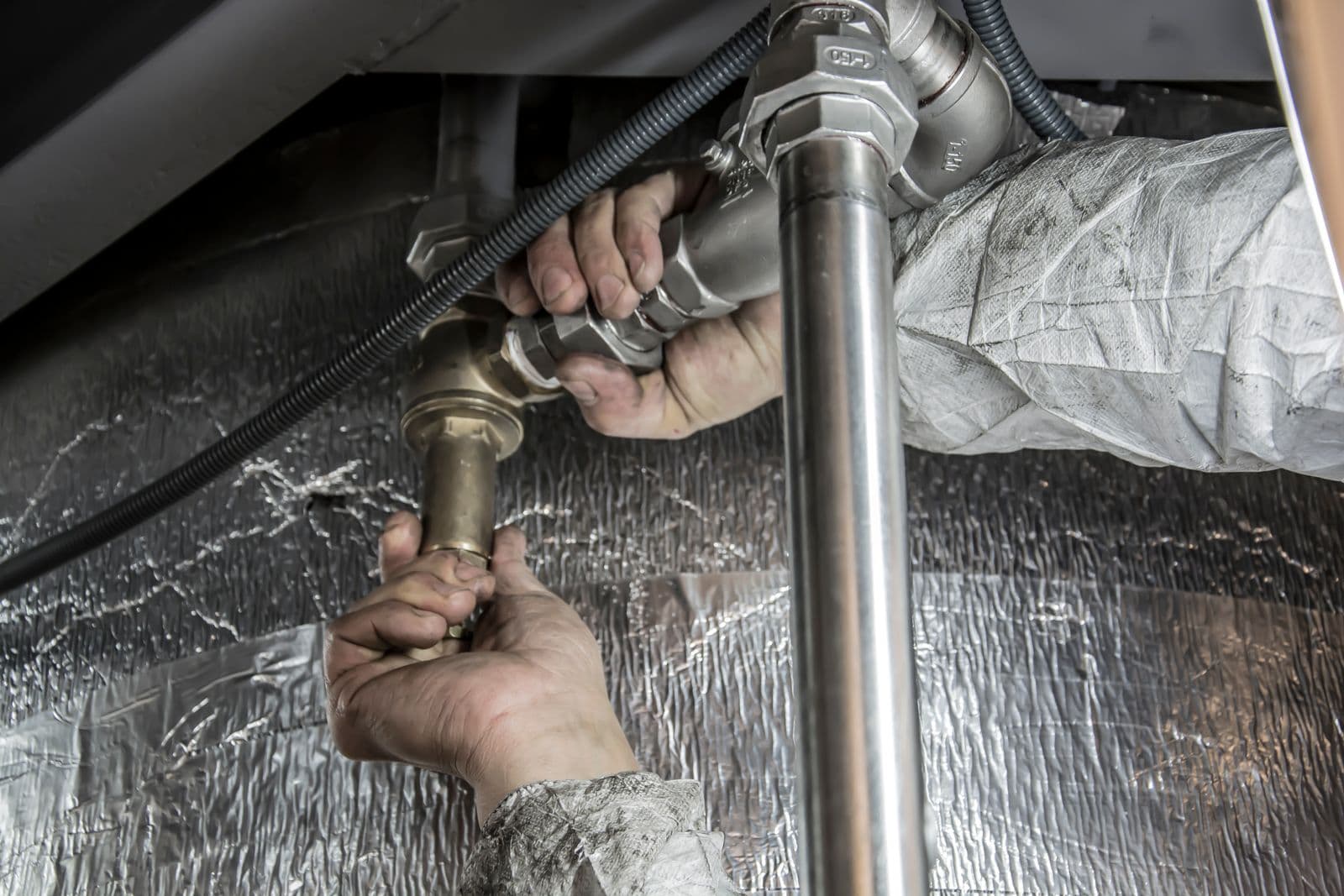 A worker's hands tightening threaded pipe fittings on supply pipework beneath equipment