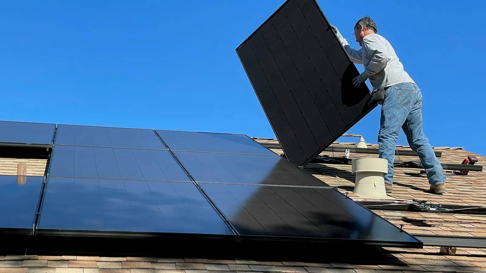 Installer lifting a black solar panel into place on a shingled home roof under a clear blue sky