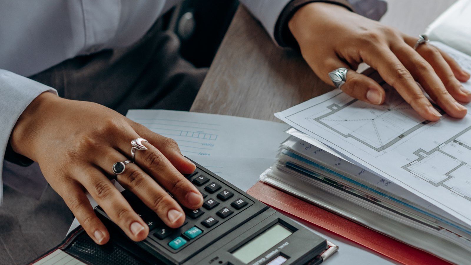 A person at a desk working through figures on a calculator, with a stack of annotated architectural drawings and a notepad beside them