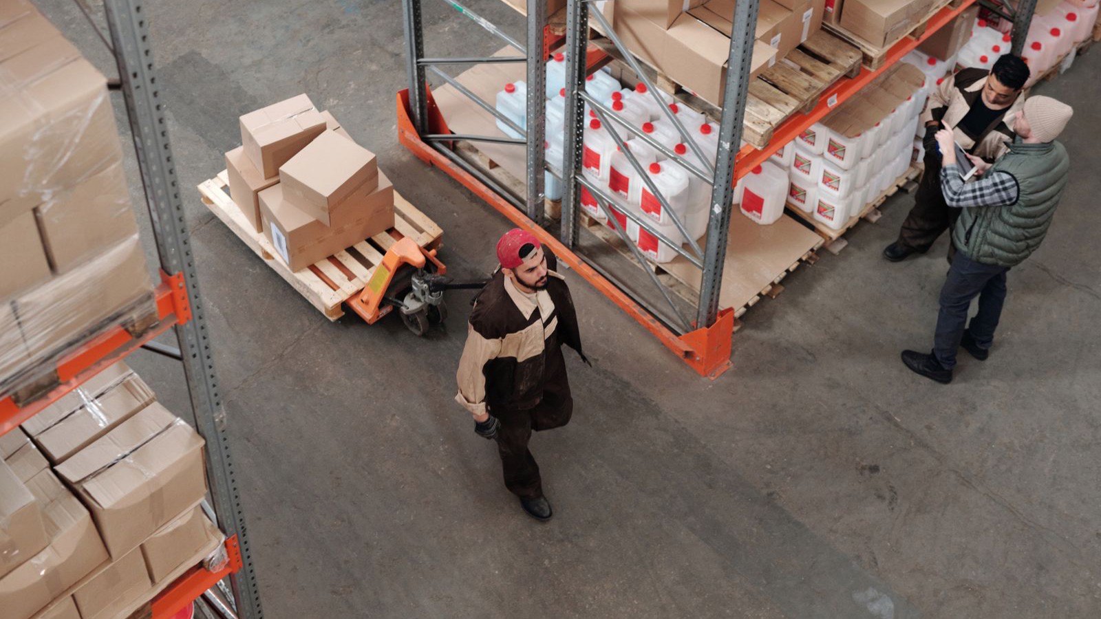 Overhead view of a warehouse aisle with stock stacked on pallets and racking, a worker moving a pallet jack, and two people reviewing inventory on a tablet